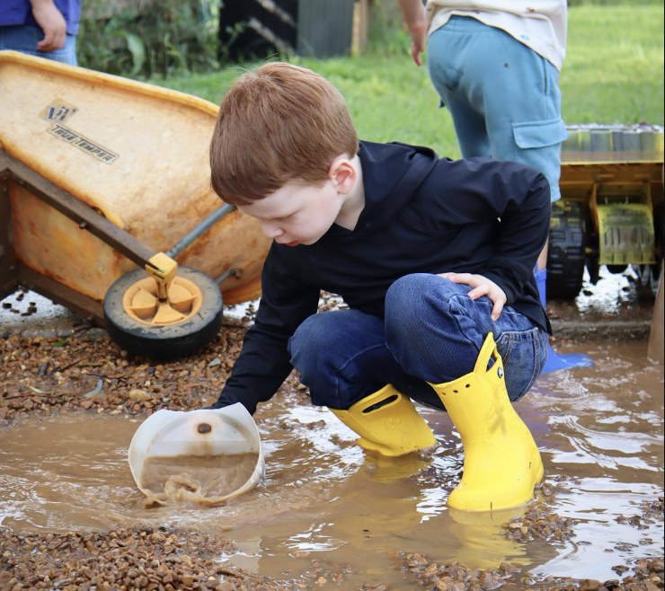 A Primary student in yellow rain boots exploring water and gravel outdoors